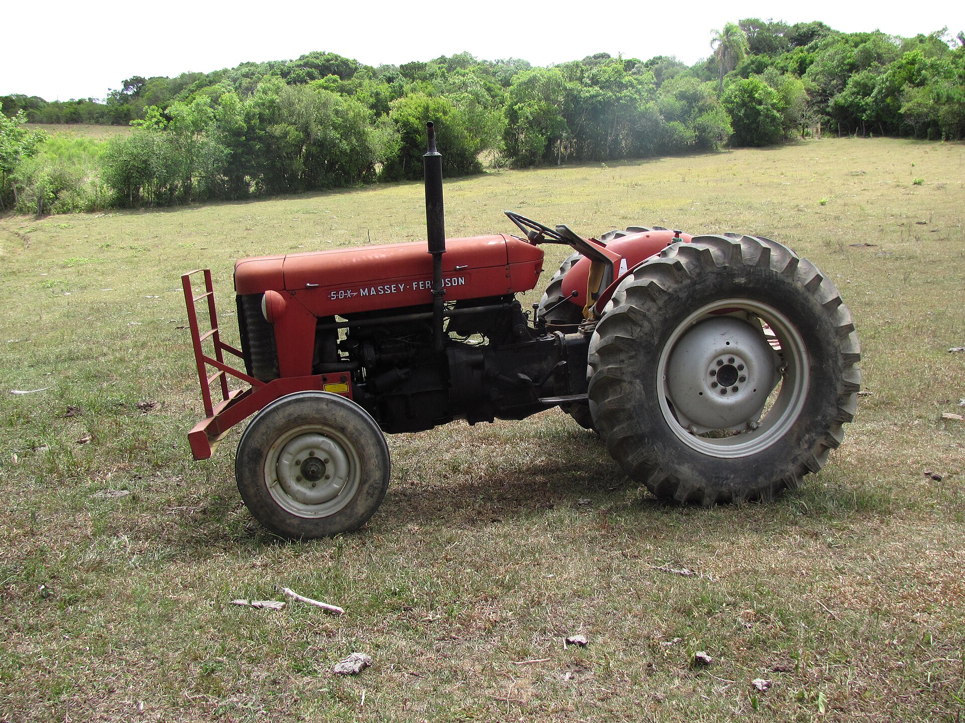 Trator Massey Ferguson 50X antigo, de pintura vermelha desbotada e pneus gastos, parado em campo de pastagem, ainda em uso na propriedade