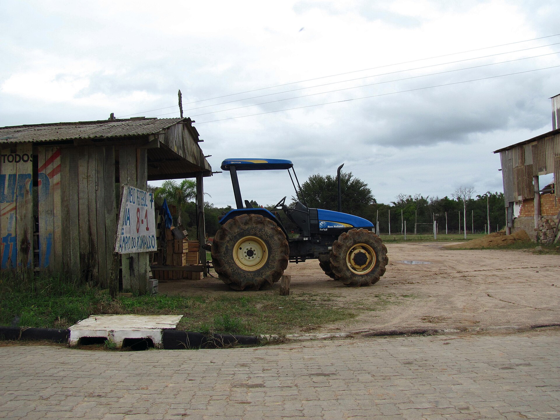 Trator New Holland TL75E azul, de porte médio e pneus enlameados, estacionado ao lado de galpão de madeira em pequena propriedade rural
