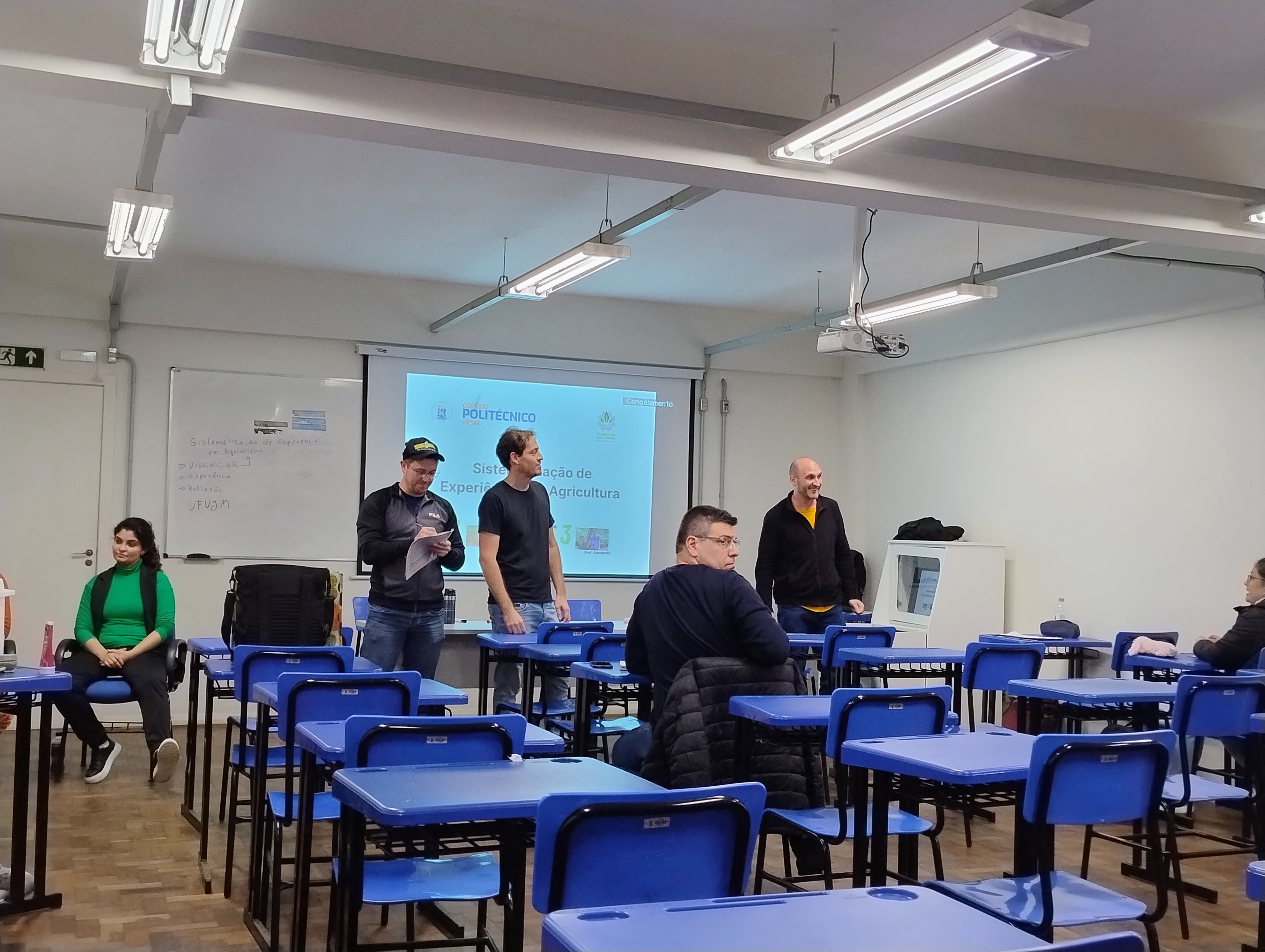 Sala de aula do Colégio Politécnico, docentes de pé apresentando o slide de abertura da disciplina de sistematização de experiências